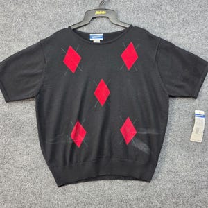 May include: A black short-sleeve knit sweater featuring a red diamond pattern with thin gray cross-hatch lines. The garment has a crew neckline and a Pendleton brand label at the inner collar, with an attached white paper retail tag on the side.