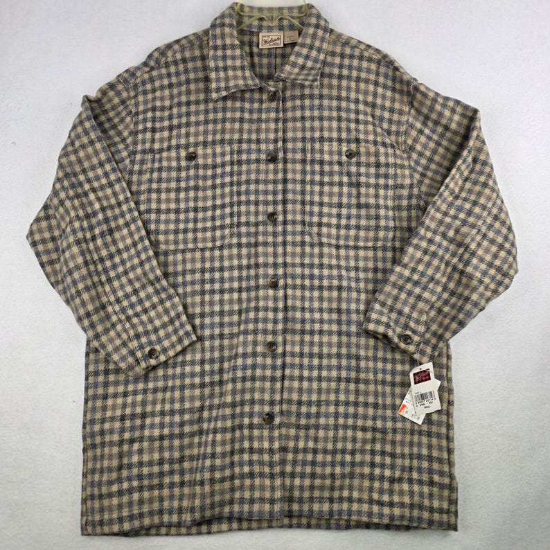 May include: A long-sleeve button-down shirt featuring a tan and dark gray plaid pattern. The shirt includes a pointed collar, two chest pockets with button closures, and buttoned cuffs. A Woolrich brand tag is visible at the neck and on a hang tag.