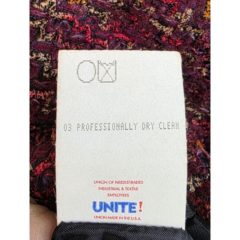 May include: Close-up of a tag with the text "03 PROFESSIONALLY DRY CLEAN" and the logo of the Union of Needletrades, Industrial & Textile Employees. The tag is against a textured, multi-colored fabric.
