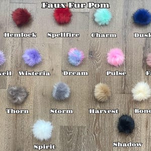 May include: A collection of faux fur pom-poms in various colors, including red, blue, pink, and purple, arranged on a wooden surface. Each pom-pom is labeled with a unique name, such as "Hemlock," "Spellfire," and "Dusk."