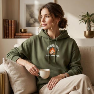 Puede incluir: Sudadera con capucha verde oliva con el texto "SUPPORT LEVEL ZEN MASTER" y un dibujo animado. La sudadera tiene una capucha con cordón y se usa con pantalones beige. Se sostiene una taza de té blanca con borde dorado.