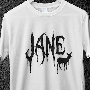 Può includere: T-shirt bianco con il nome "JANE" in un font gotico gocciolante e una silhouette nera di cervo. La maglietta è appesa a una gruccia bianca su uno sfondo scuro strutturato.