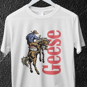 Può includere: T-shirt bianca con un cowboy che cavalca un cavallo imbizzarrito, con la parola "Geese" in testo verticale rosso. Il cowboy indossa un cappello e una camicia blu. La maglietta è appesa a una gruccia bianca.