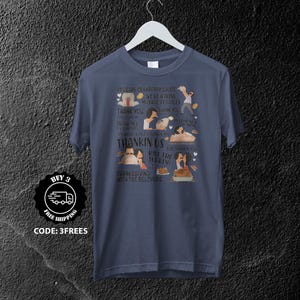 Può includere: T-shirt blu navy con grafica a tema Ringraziamento con personaggi dei cartoni animati e testo. Il testo include frasi come "Passa la salsa di mirtilli rossi". La maglietta è su un appendiabiti.