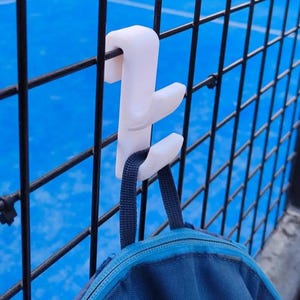 May include: A white plastic hook is attached to a black metal fence, holding a blue backpack. The hook has a unique design with multiple prongs. The background features a blue surface, possibly a sports court.