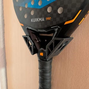May include: A black and blue Kuikma 990 paddleball racket with a black grip. The racket has a checkered pattern and a blue design. The grip is wrapped in black tape with the Alpha logo.
