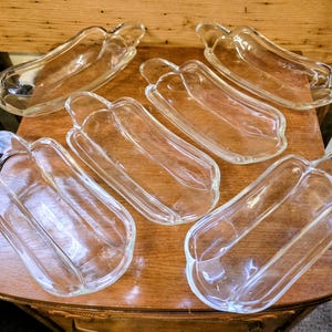 Puede incluir: Seis fuentes de vidrio transparente, con forma de perritos calientes, están dispuestas sobre una mesa de madera. Cada fuente tiene un asa y un diseño curvo, ideal para servir aperitivos. Las fuentes son transparentes.