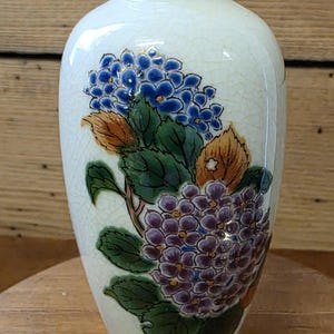 Op de afbeelding: Crèmekleurige keramische vaas met een bloemenontwerp van blauwe en paarse hortensia's, groene bladeren en oranje accenten. De vaas heeft een craquelé glazuur en een gouden rand, wat een klassieke decoratieve stijl laat zien.