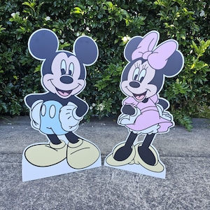 May include: Two cartoon character cutouts, Mickey Mouse and Minnie Mouse, stand side-by-side. Mickey is in black, white, and light blue, while Minnie is in black, pink, and white. Both have yellow shoes and are smiling. They are standing on a concrete surface.