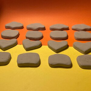 May include: A collection of small, light brown clay shapes, including irregular forms, hearts, and squares, are arranged on a yellow surface against an orange backdrop. The shapes are uniform in colour and texture.