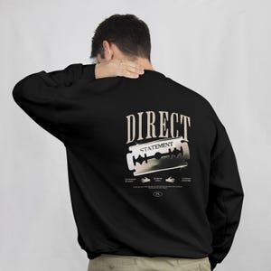 Puede incluir: Sudadera negra de manga larga con un gráfico en la espalda. El gráfico presenta las palabras "DIRECT STATEMENT" sobre una ilustración de una cuchilla de afeitar. Texto adicional e ilustraciones de manos debajo.