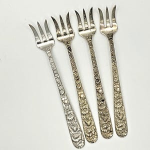 May include: Four silver-toned metal cocktail forks arranged in a row. Each fork features a three-tined head and a long, slender handle decorated with an intricate, embossed floral pattern that extends from the base to the neck of the fork.
