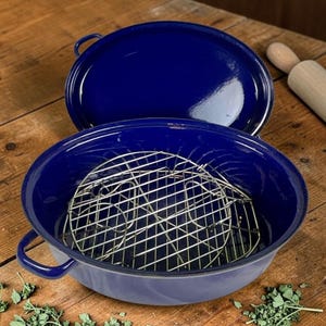 May include: A deep, round, dark blue enameled roasting pan with two side handles and a matching lid. Inside the pan sits a removable circular metal wire rack. The exterior and interior surfaces have a smooth, glossy finish in a solid navy blue.