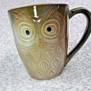 May include: A ceramic coffee mug with an owl design. The mug features a detailed owl face with large eyes and feather-like patterns. The color palette includes shades of brown, beige, and green, with a dark handle.