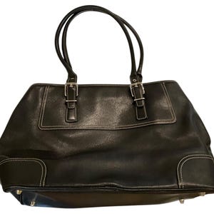 May include: A black tote bag featuring two shoulder straps, silver-toned metal buckles at the base of the handles, and white contrast stitching along the edges. The bag has a structured rectangular base with small silver-toned metal feet.