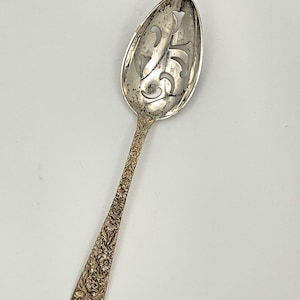 May include: A silver-toned serving spoon featuring an oval bowl with intricate cut-out scrollwork patterns. The handle is decorated with a detailed embossed floral motif that extends from the base of the bowl to the end of the handle.