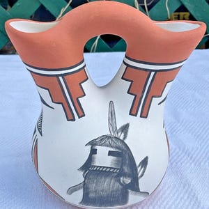 May include: A handcrafted Native American pottery vessel with a distinctive double-spout design. The vessel features a cream-colored body adorned with geometric patterns in black and orange. A central figure is depicted in black. The top spouts are orange.