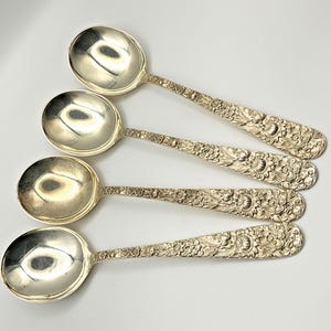 May include: A set of four silver-toned spoons with polished, rounded bowls and handles featuring an intricate, embossed floral pattern. The metal surfaces exhibit a reflective finish, and the handles are decorated with detailed flower motifs.