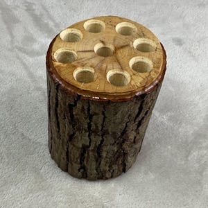May include: A cylindrical wooden pencil holder made from a tree trunk section. The top has nine evenly spaced holes. The exterior bark is rough and brown, while the interior is a lighter wood color. The holder is approximately 4 inches tall.