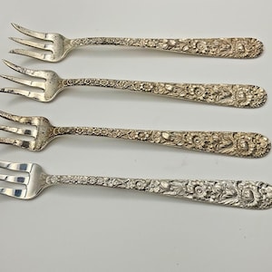 May include: A set of four silver-toned metal cocktail forks arranged horizontally. Each fork features three tines and a handle decorated with an intricate, embossed floral pattern that covers the entire length of the handle surface.
