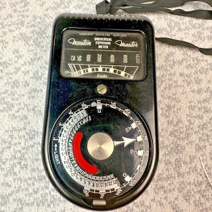 May include: A vintage Weston Master Universal Exposure Meter, black with a circular dial and a needle indicator. The meter has a black strap and features the text "Weston Master" and "Universal Exposure Meter". The dial includes numbers and a red arc.