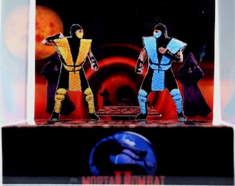 Op de afbeelding: Een Mortal Kombat II diorama met twee personages in vechthouding tegen een rode en zwarte achtergrond. Eén personage is geel en zwart, de ander blauw en zwart. Het Mortal Kombat II logo staat onderaan.