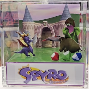 Puede incluir: Diorama de Spyro the Dragon en un cubo transparente. Presenta un dragón morado, una criatura verde y un escenario de castillo. La base es verde con gemas de colores. El logotipo de Spyro se muestra en la parte inferior.