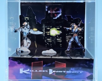 Puede incluir: Una vitrina de acrílico transparente con un diorama de dos figuras en un entorno urbano. Las figuras son del videojuego "Killer Instinct". La base de la vitrina tiene el logotipo del juego en texto blanco sobre un fondo morado y negro.