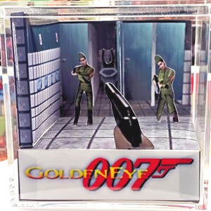 May include: A miniature diorama of a scene from GoldenEye 007. The scene shows a bathroom setting with paper cutouts of characters in green uniforms. The GoldenEye 007 logo is displayed on the base of the diorama.