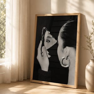 May include: Monochrome framed print showing a woman holding a shard of mirror, reflecting her eye. The artwork is in a wooden frame and leans against a wall. A vase with a plant and a window with curtains are in the background.