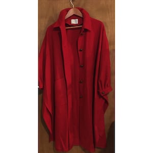 Op de afbeelding: Een lange, rode jas met een kraag en een knoopsluiting. De jas heeft een cape-achtig ontwerp met wijde mouwen en een lengte tot halverwege de kuit. Hij hangt aan een houten kleerhanger tegen een bruine houten achtergrond.