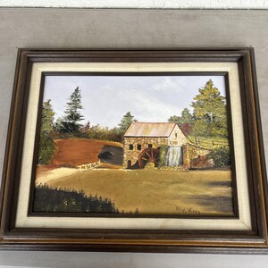 May include: Oil painting of a rural landscape featuring a stone mill with a water wheel and waterfall. The artwork is framed in a dark brown wooden frame with a cream-colored matting. The scene includes trees, a grassy field, and a cloudy sky.