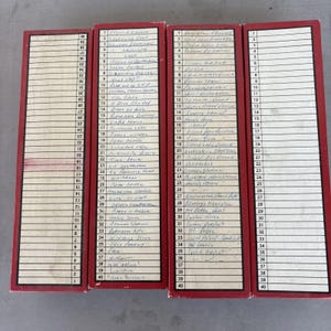 May include: Four vintage index card holders with red borders and white cards filled with handwritten notes. The cards are organized in rows and columns, with each card containing a list of names and places. The holders are stacked together.