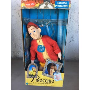 May include: A Talking Pinocchio doll in a cardboard box. The doll has a plastic head, blue eyes, and wears a red jacket over a yellow shirt and shorts. The box features the text "TRY ME!", "TALKING PINOCCHIO", and "THE ADVENTURES OF PINOCCHIO".