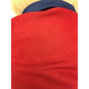 May include: Close-up of a red fabric with horizontal ribbed texture. A navy blue collar is visible at the top, and a tan fabric is partially visible above the collar. The overall impression is of a garment.