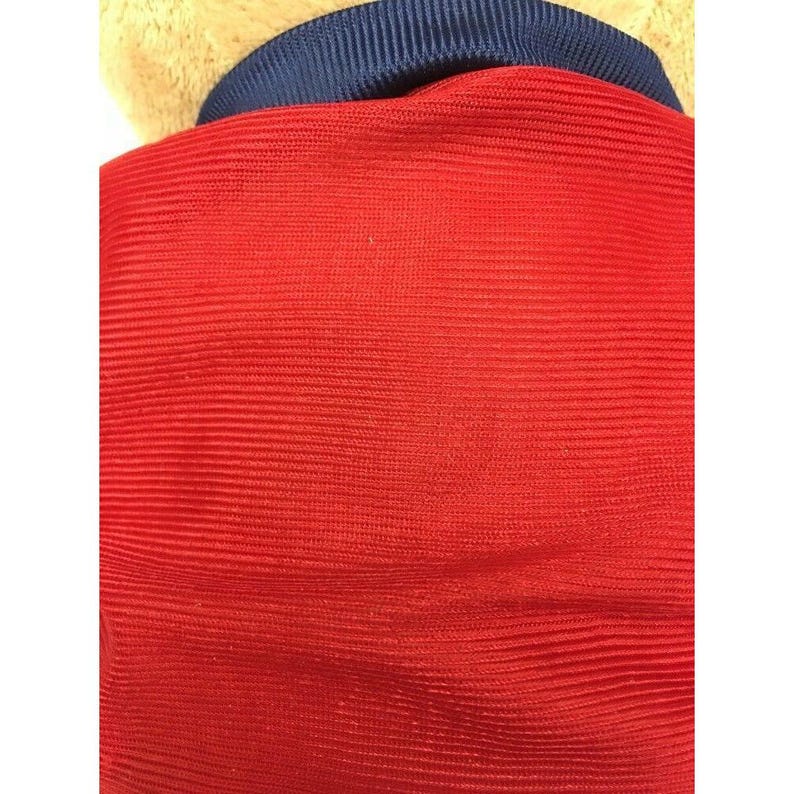 May include: Close-up of a red fabric with horizontal ribbed texture. A navy blue collar is visible at the top, and a tan fabric is partially visible above the collar. The overall impression is of a garment.