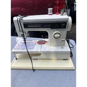 Pode incluir: Uma máquina de costura Kenmore 10 branca com painel de controle preto para seleção de pontos, um seletor circular para comprimento do ponto, um suporte de carretel superior e um adesivo em formato de tomate na base plana.