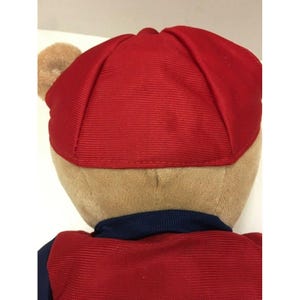 May include: A plush teddy bear wearing a red cap and a red and navy blue outfit. The bear's fur is tan, and the cap is a solid red color. The outfit has a navy blue collar.