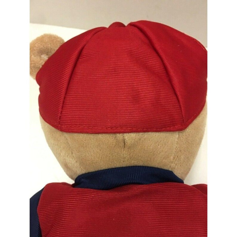 May include: A plush teddy bear wearing a red cap and a red and navy blue outfit. The bear's fur is tan, and the cap is a solid red color. The outfit has a navy blue collar.