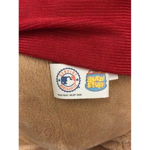 May include: Close-up of a red fabric item with a tan plush material underneath. A tag with the MLB logo and the words "Genuine Merchandise" is visible, along with a "Good Stuff" tag. The tags read "Good Stuff / MLB 2009".