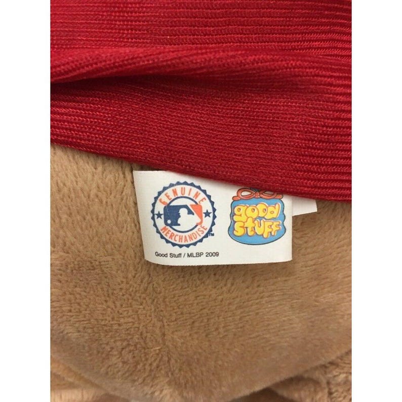 May include: Close-up of a red fabric item with a tan plush material underneath. A tag with the MLB logo and the words "Genuine Merchandise" is visible, along with a "Good Stuff" tag. The tags read "Good Stuff / MLB 2009".
