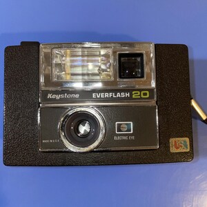 May include: A vintage Keystone Everflash 20 camera. The black camera has a silver lens, a clear flash, and the words "Keystone" and "EVERFLASH 20" printed on the front. The words "ELECTRIC EYE" and "MADE IN U.S.A." are also visible.