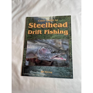 Könnte beinhalten: Ein Taschenbuch mit dem Titel Color Guide to Steelhead Drift Fishing von Bill Herzog. Das Cover zeigt ein Foto eines Steelhead-Fisches mit einem rosa Köder im Maul vor dem Hintergrund eines Waldes und fließendem Flusswasser.