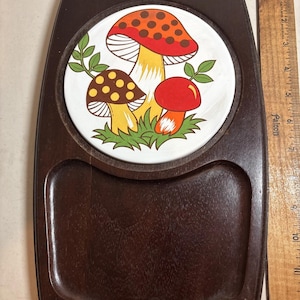 May include: A vintage wooden serving tray with a circular ceramic tile featuring a colorful mushroom design. The tray is dark brown with a rectangular shape, a handle, and a recessed area. A wooden ruler is on the right side.
