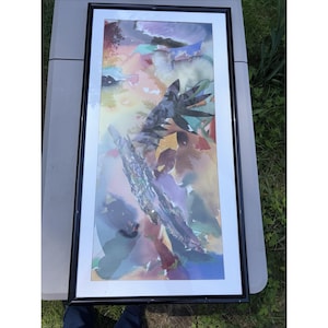 May include: Abstract watercolor painting in a black frame. The artwork features a vibrant mix of colors, including blues, purples, greens, and yellows. The composition includes layered shapes and textures, creating a dynamic and expressive piece. The frame is rectangular.