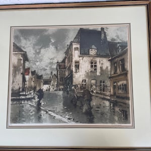 Op de afbeelding: Ingelijste prent van een Europese straatscène in gedempte tinten. Het kunstwerk toont gebouwen langs een natte straat met figuren en paardenkoetsen. De lucht is bewolkt en de prent zit in een gouden lijst.