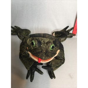 May include: A plush frog toy with dark green mottled skin, large bright green eyes, and a protruding red tongue. The stuffed animal features four splayed limbs with detailed toes and is positioned next to a red ruler for scale reference.