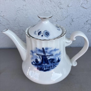 May include: White ceramic teapot featuring a blue and white windmill scene. The teapot has a lid with floral designs and a silver trim. The handle and spout are curved. A classic design for tea lovers.