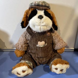 May include: A plush St. Bernard dog wearing a brown corduroy overall, a knitted sweater, and a plaid cap. The dog has brown and black fur with a white muzzle and paws. A heart-shaped patch is on the overall pocket.