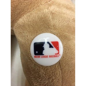May include: A white, circular Major League Baseball logo pin with a black, red, and white design. The pin is affixed to a plush, tan-colored surface.